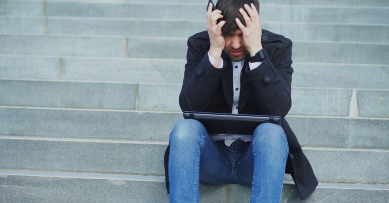 A worried man sitting on steps with a laptop while holding his phone